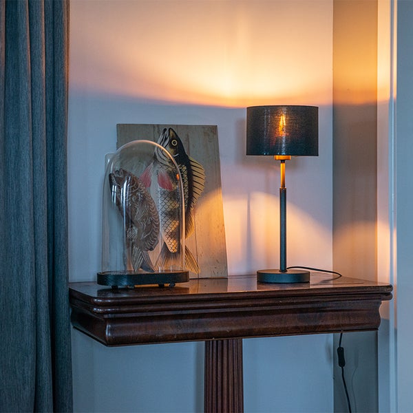 Hell leuchtende Tischleuchte mit dunklem Stoffschirm und Metallfuß auf einem Holztisch neben Fisch-Dekorationen und einem Vorhang.