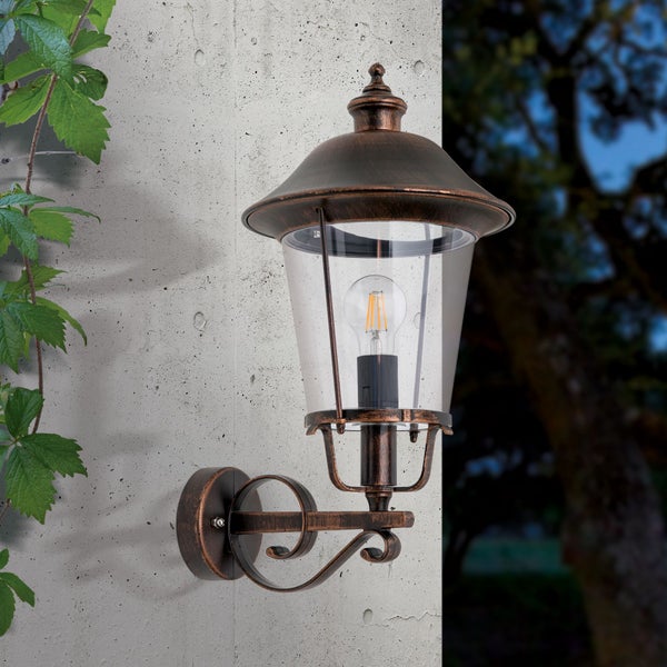 Klassische Außenwandleuchte in brauner Antik-Optik aus Metall und Glas mit Filament-Leuchtmittel an einer Betonwand im Außenbereich neben Blättern.