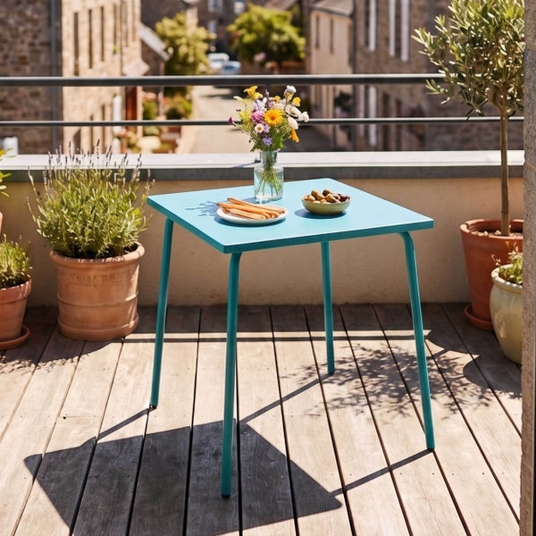 Quadratischer Bistro-Gartentisch aus Metall auf einem hellen, sonnigen Balkon mit Holzdeck, Pflanzen in Tontöpfen und Blumen auf dem Tisch.
