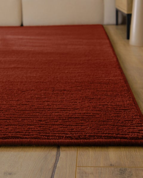 Strukturierter Teppich mit Rippenmuster in einem hellen Wohnzimmer mit Boden in Holzoptik, hellem Sofa, Holzhocker und offenem Buch.