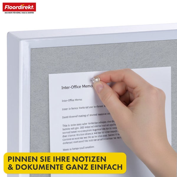 Person befestigt eine Notiz mit einer Reißzwecke an einer Pinnwand mit weißem Rahmen. Floordirekt Logo.