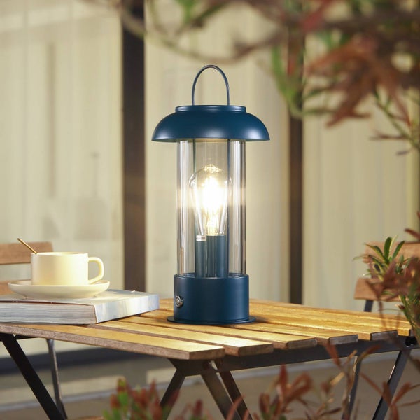 Blaue, tragbare Außenlampe mit Glühbirne auf einem hölzernen Gartentisch neben einer weißen Kaffeetasse und einem Buch in einer hellen Umgebung.