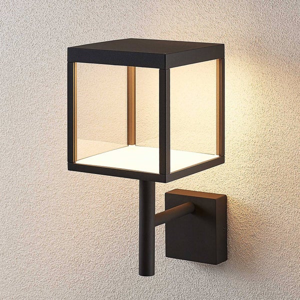 Moderne Außenwandleuchte in Würfelform mit schwarzem Metallgehäuse und klaren Glasflächen, montiert an einer Wand mit warmem Licht.