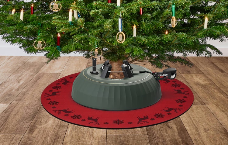 Krinner Christbaumständer auf roter Weihnachtsbaumdecke mit Rentier-Muster unter einem geschmückten Weihnachtsbaum auf Holzboden.