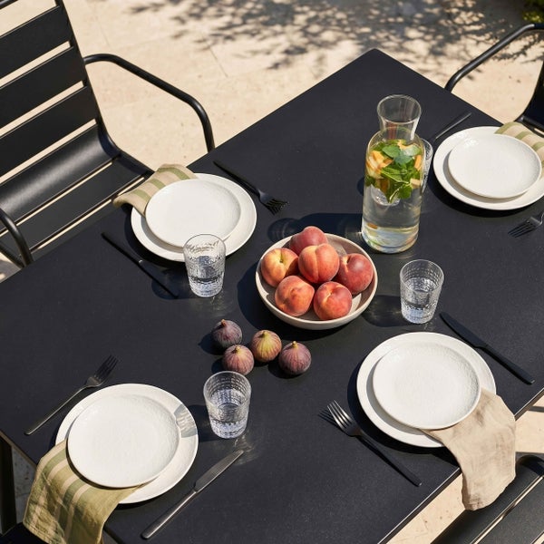 Gedeckter Gartentisch aus Metall mit Tellern, Gläsern, Besteck, Wasserkaraffe und Obstschale in heller Außenumgebung.