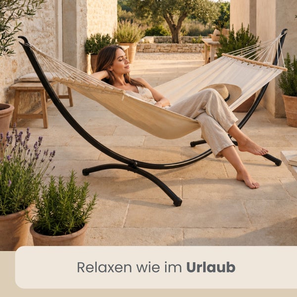 Eine Frau entspannt in einer cremefarbenen Hängematte mit schwarzem Metallgestell auf einer hellen Terrasse mit Steinboden und Topfpflanzen.