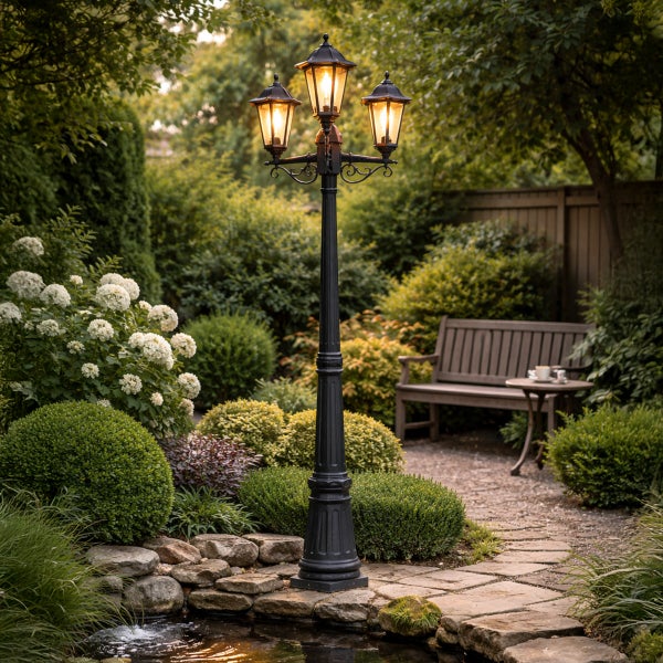 Dreiflammige Gartenlaterne aus Metall in einer Gartenanlage mit Teich, Steinpfad, Holzbank und blühenden Pflanzen.