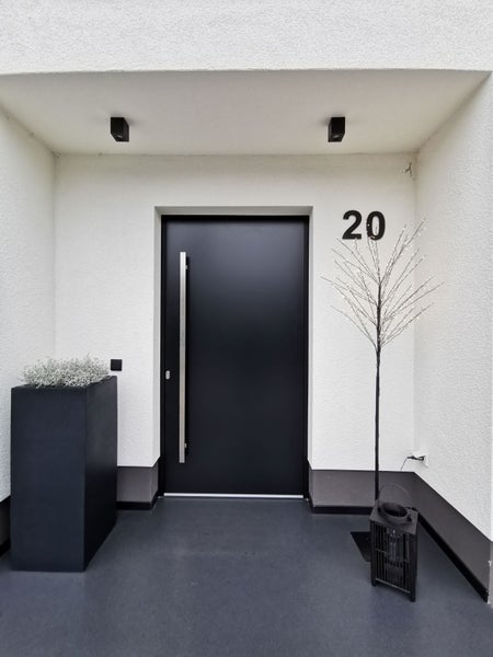 Moderner Eingangsbereich mit schwarzer Haustür, langem silberfarbenem Griff, Hausnummer 20, Pflanzkübel und Lichterbaum.