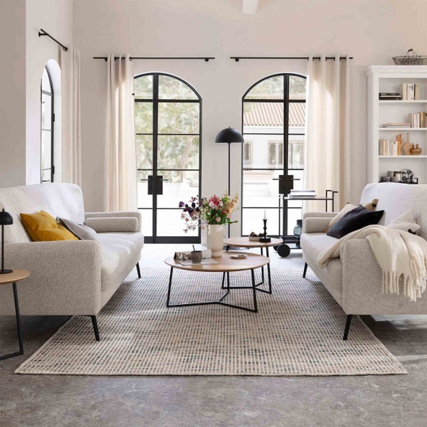 Helles Wohnzimmer mit zwei beigen Sofas, zwei runden Couchtischen aus Holz, einem gemusterten Teppich und großen Rundbogenfenstern.