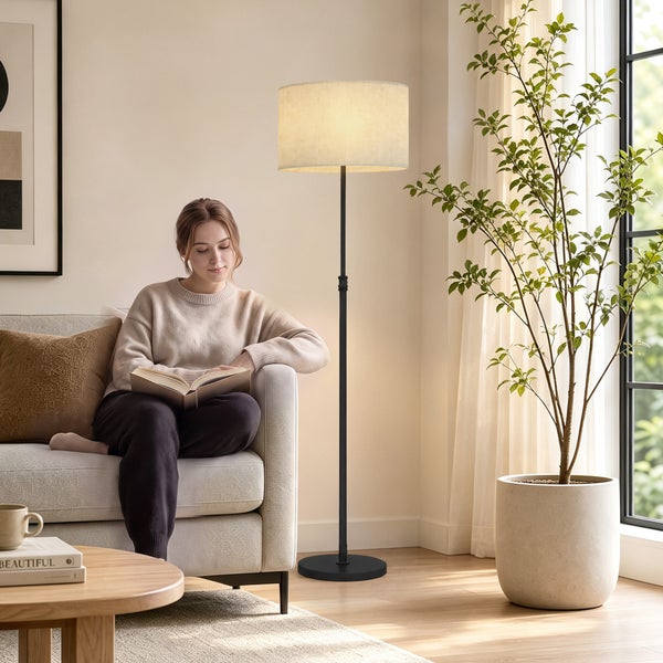 Eine Frau liest ein Buch auf einem Sofa neben einer Stehleuchte mit einem zylindrischen Lampenschirm in einem hellen, modern eingerichteten Wohnraum.