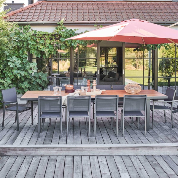 Modernes Gartenmöbel-Set auf einer Holzterrasse mit großem Holztisch, Metallstühlen und einem Sonnenschirm vor einer Fensterfront.