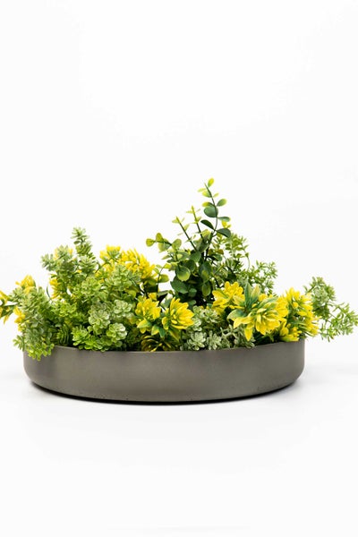 Künstliches Pflanzengesteck mit gelben Blüten und grünem Blattwerk in einer runden grauen Dekoschale.