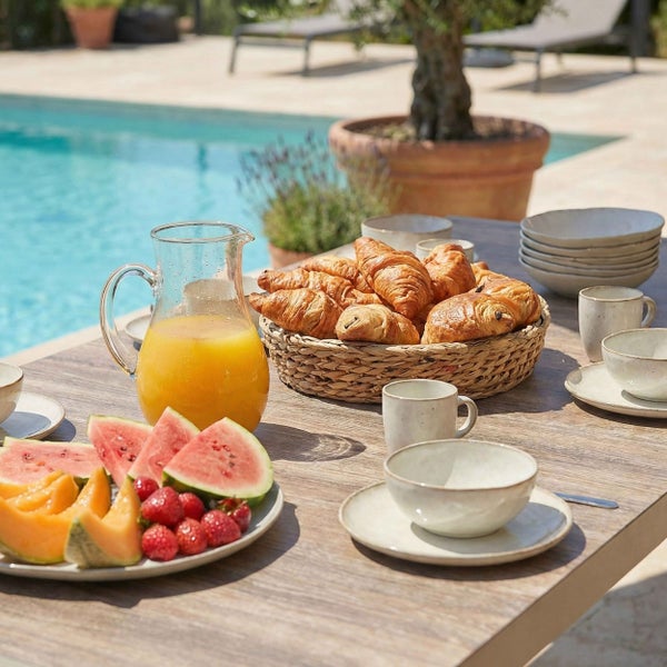 Gedeckter Holztisch im Freien am Pool mit Keramikgeschirr, Fruchtsaft in einer Glaskaraffe, Gebäck im Flechtkorb und frischem Obst.