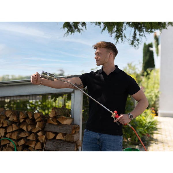 Ein Mann bedient einen Gasbrenner im Garten neben einem Holzstapel.
