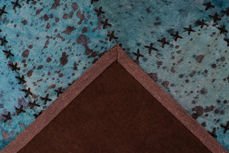 Patchwork-Teppich aus Kuhfell in Blau mit dunklen Flecken, dekorativen Kreuzstichen und brauner Filzrückseite.