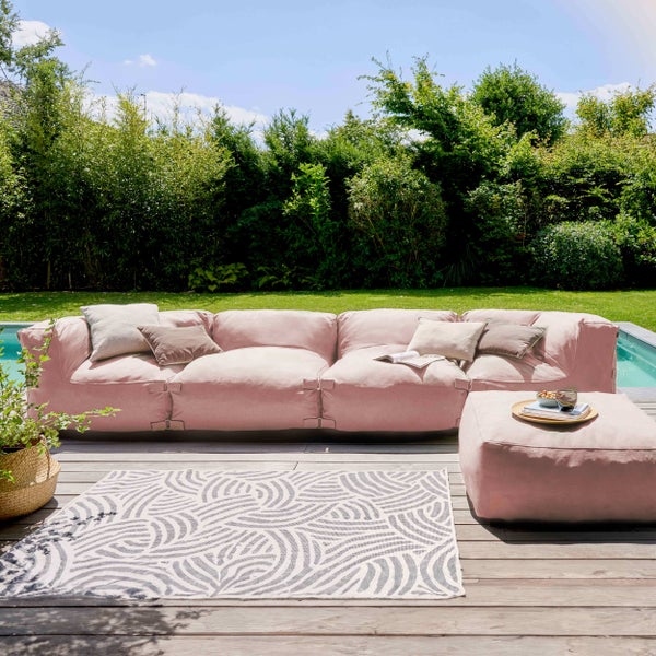 Modernes Lounge-Set mit rosa Sofa, Pouf und gemustertem Außenteppich auf einer Holzterrasse am Pool im Garten.