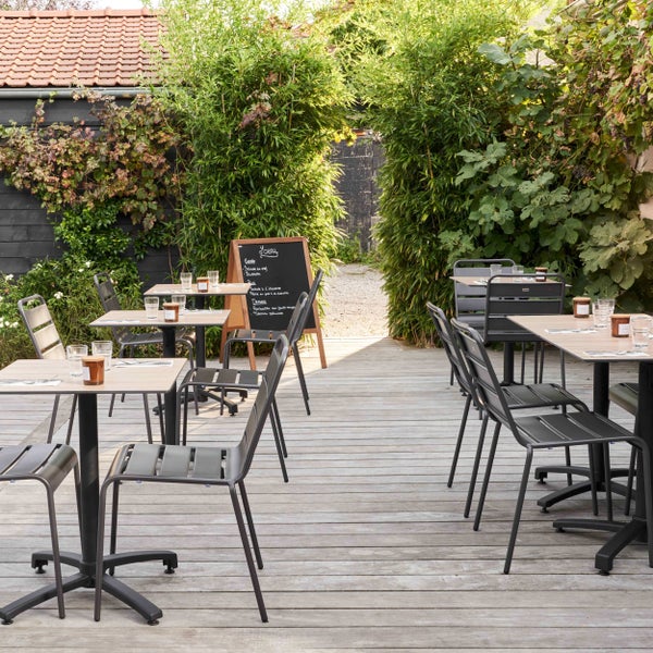 Helle Außenterrasse mit Holzdielen, modernen schwarzen Metallstühlen und Tischen mit Holzplatte, umgeben von dichtem Grün und Bambuspflanzen.