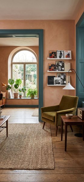Gemütliches Wohnzimmer mit terracottafarbenen Wänden, Samtsessel in Olivgrün, Jute-Teppich und weißen Wandregalen für Zeitschriften.