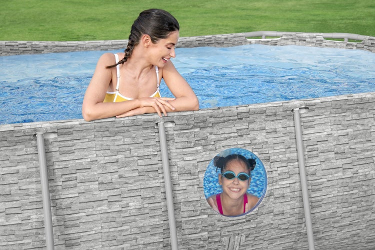 Aufstellpool mit Außenwand in Steinoptik, eine Frau lehnt am Beckenrand und ein Kind blickt durch ein integriertes Sichtfenster im Wasser.