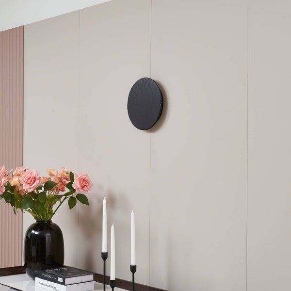 Runder schwarzer Wandhaken mit strukturierter Oberfläche an einer Paneelwand über einer Vase mit rosa Rosen und weißen Kerzen.