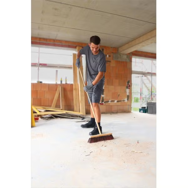 Ein Handwerker kehrt mit einem Holzbesen den Staub von einem Estrichboden auf einer Baustelle.