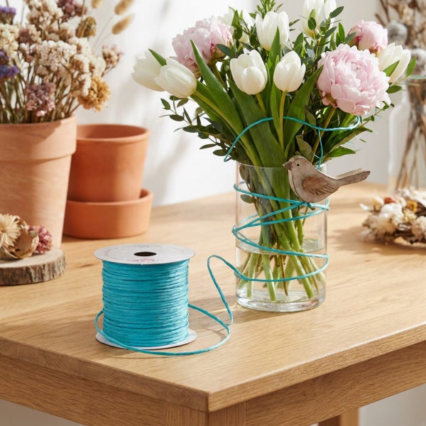 Rolle mit dekorativer Kordel auf einem Holztisch neben einer Glasvase mit Blumen und einem kleinen Vogel-Dekorationselement.