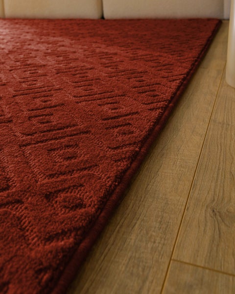 Roter Teppich mit geometrischer Struktur und eingefasstem Rand auf Boden in Holzoptik.
