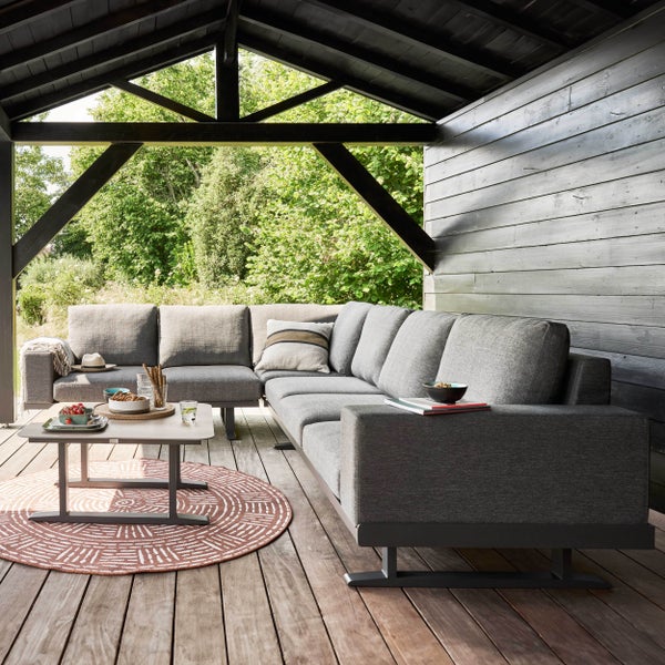 Große graue L-förmige Lounge-Sitzgruppe auf einer Holzterrasse unter einer Überdachung mit Loungetisch und Teppich vor einem Garten.