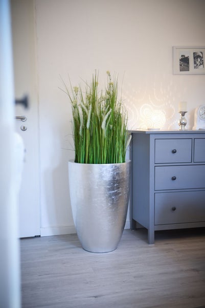 Hoher silberfarbener Pflanzkübel mit künstlichem Gras in einem hellen Zimmer neben einer grauen Kommode.