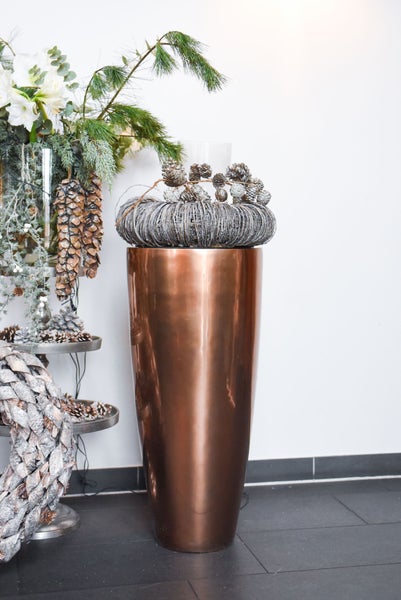 Hohe konische Bodenvase in Kupferoptik mit festlicher Winterdekoration aus Kränzen und Tannenzapfen in einem hellen Raum.