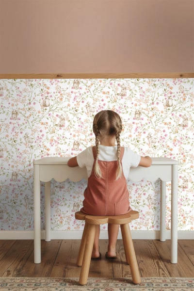 Mädchen sitzt an einem Tisch vor einer Tapete mit Blumen- und Hasenmuster.
