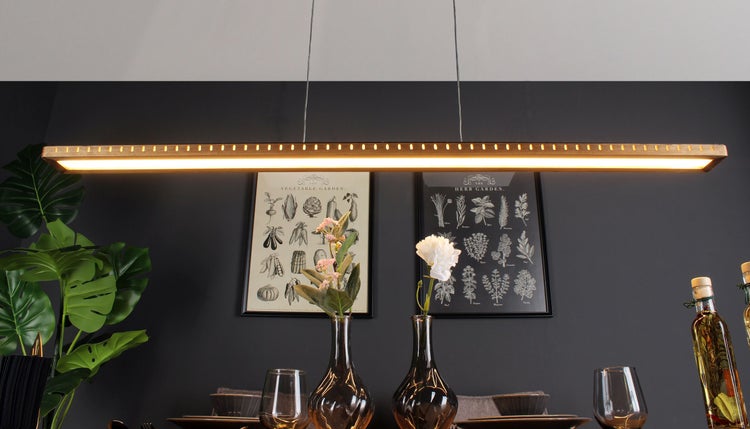 Moderne hölzerne LED-Pendelleuchte über einem Esstisch in einem Zimmer mit botanischen Bildern an einer dunklen Wand.