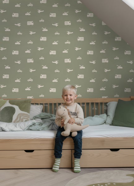 Kinderzimmer mit Tapete im Fahrzeugmuster, Holzbett mit Schubladen und sitzendem Kind mit Stoffdinosaurier.