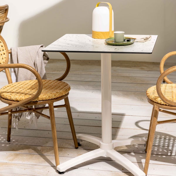 Moderner quadratischer Bistrotisch mit weißem Gestell, Terrazzo-Platte, Laterne und zwei Rattanstühlen auf einer Holzterrasse.
