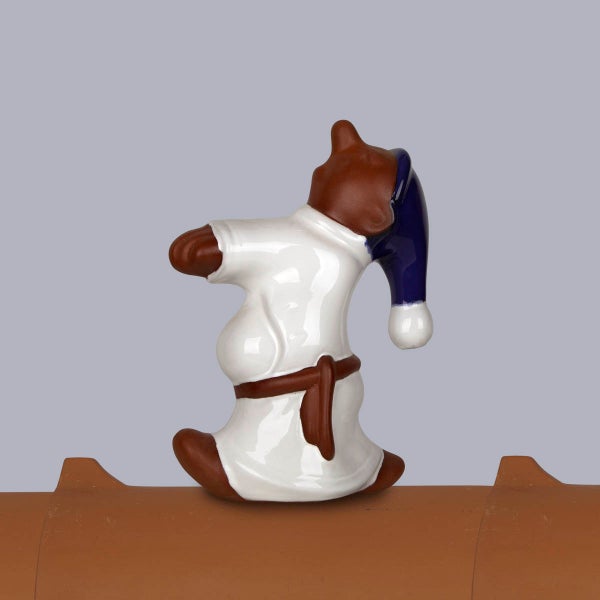 Firstfigur aus Keramik in Form eines braunen Bären mit weißem Nachthemd und blauer Schlafmütze auf einem Firstziegel.