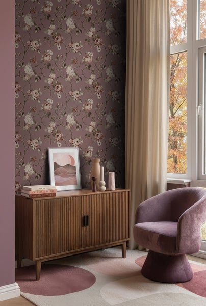 Vliestapete mit floralem Rosenmuster an einer Wand im Wohnzimmer neben einem Holzsideboard und einem Sessel.
