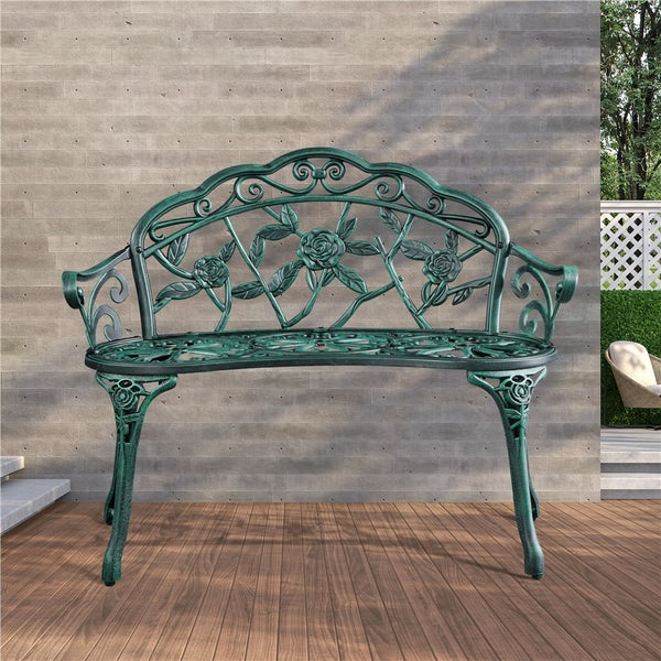 Verzierte Gartenbank aus Metall mit Rosen-Ornamenten auf einer Holzterrasse vor einer Steinwand in einem hellen Außenbereich.