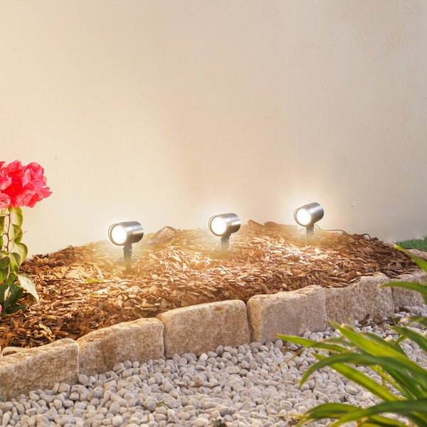Drei Gartenstrahler aus Edelstahl im Rindenmulchbeet beleuchten eine Wand, eingefasst von Natursteinen und weißen Kieseln.