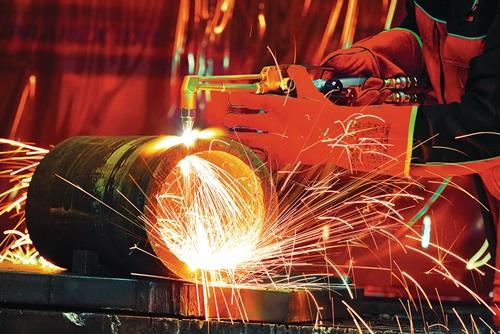 Eine Person in Schutzkleidung und Schweißerhandschuhen schneidet ein Metallrohr mit einem Schneidbrenner unter Funkenflug.