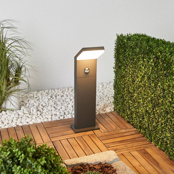 Moderne anthrazitfarbene Außenstandleuchte mit Bewegungsmelder und LED auf einer Holzterrasse neben einer Hecke und Kies.