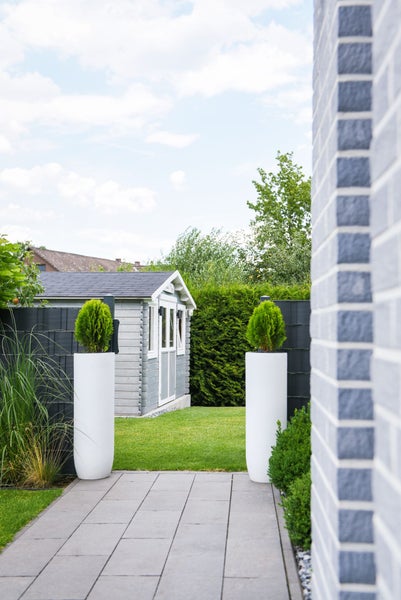 Moderner Garten mit grauem Gartenhaus, Plattenweg, zwei weißen Pflanzkübeln und Sichtschutzzaun auf gepflegtem Rasen.