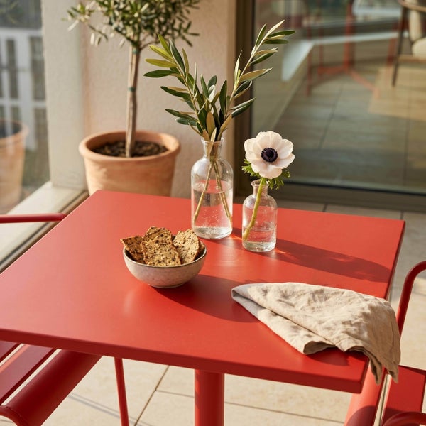 Quadratischer Gartentisch aus rotem Metall auf einem hellen Balkon mit passenden Stühlen, dekoriert mit Vasen, Crackern und einer Stoffserviette.