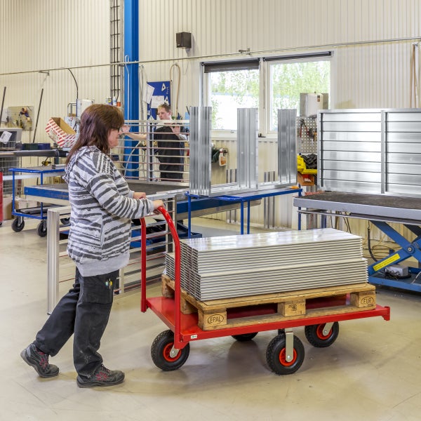 Eine Frau schiebt einen roten Wagen mit einer Palette Aluminiumprofile in einer Werkshalle.