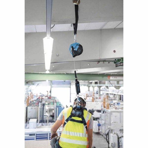 Person mit Zarges Auffanggurt und Warnweste, gesichert an einem Höhensicherungsgerät an einer Deckenschiene in einer Industriehalle.