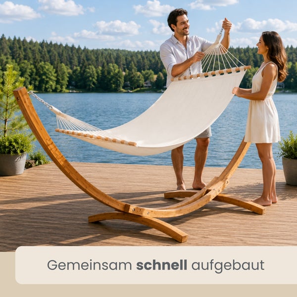 Ein Paar baut eine Hängematte aus Stoff mit einem geschwungenen Holzgestell auf einer Holzterrasse an einem See auf.