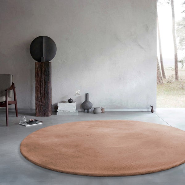 Runder Teppich in Terracotta mit flauschiger Oberfläche in einem minimalistischen Wohnzimmer mit Betonwand und Blick ins Grüne.