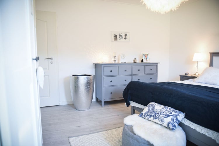 Modernes, helles Schlafzimmer mit grauer Kommode, silbernem Pflanzgefäß, Bett mit blauer Decke und hellem Laminatboden.