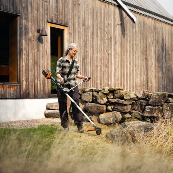 Frau arbeitet mit einem Akku-Rasentrimmer im Garten vor einem Holzhaus.