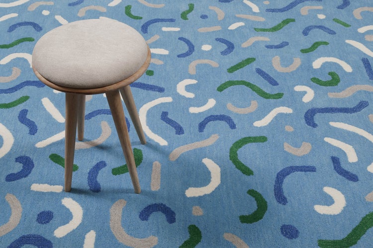 Runder Hocker mit beigem Polstersitz und Holzbeinen auf einem blauen Teppich mit abstrakten Wellenmustern in verschiedenen Farben.