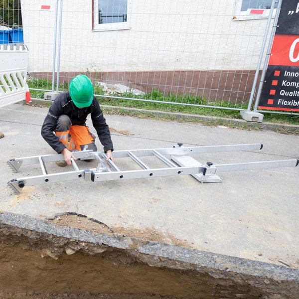 Bauarbeiter mit grünem Helm montiert eine silberne Aluminiumleiter auf einer Baustelle neben einem Graben.